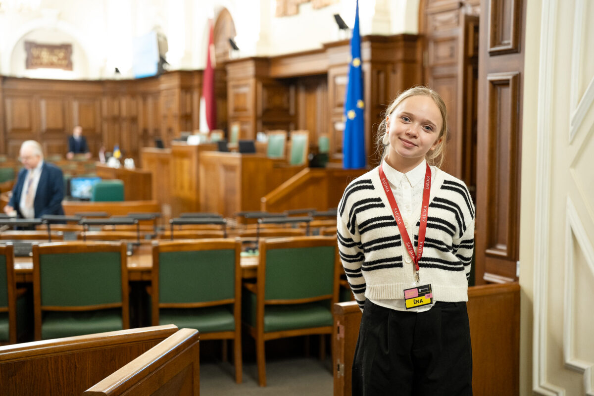 Smaidīga meitene ar sarkanu kakla lenti ar uzrakstu SAEIMA un piespraudi ĒNA stāv Saeimas sēžu zālē uz deputātu solu un Eiropas Savienības karoga fona.