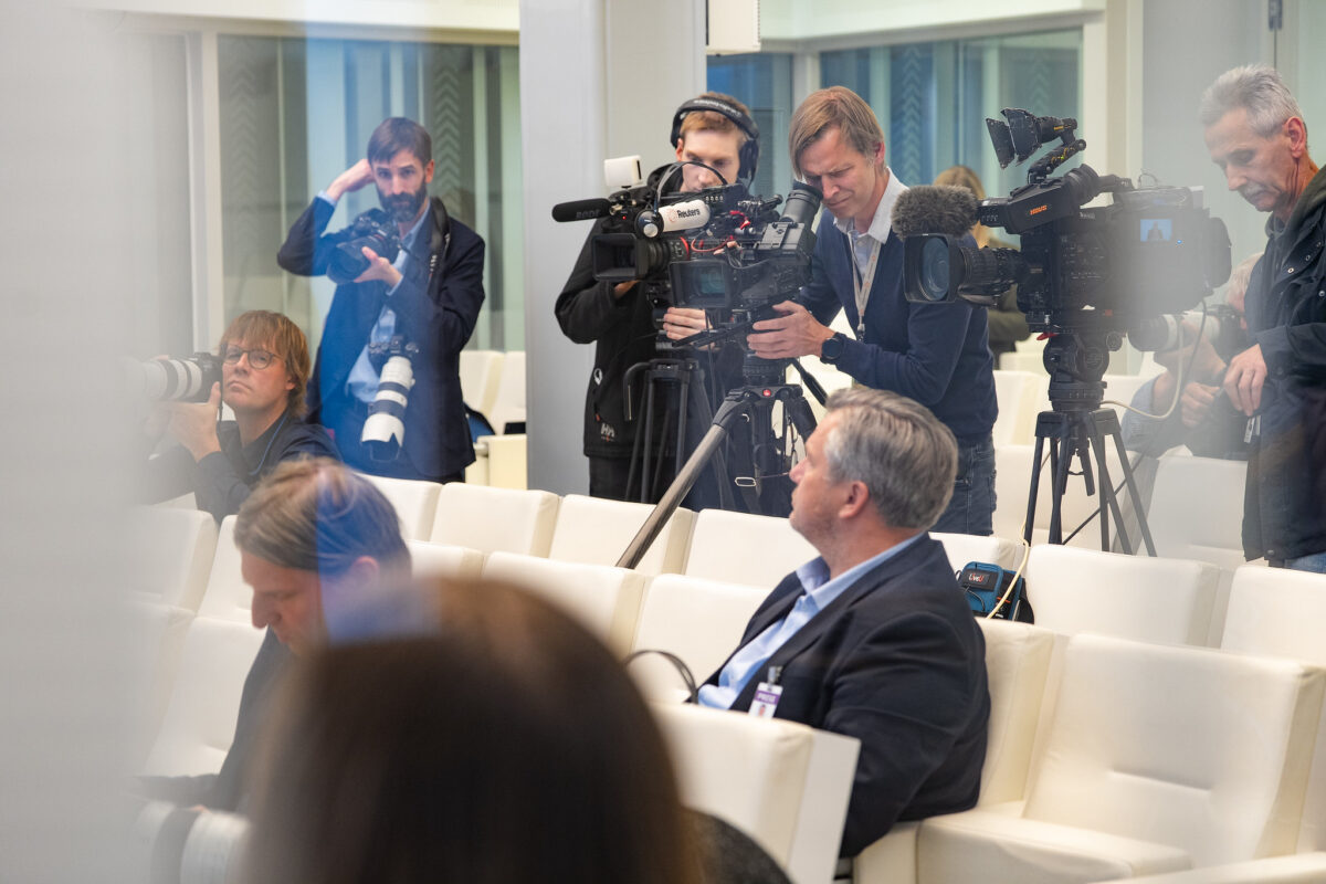 Operatori un fotogrāfi ar profesionālu filmēšanas tehniku un kamerām dokumentē pasākumu konferenču telpā ar baltiem krēsliem.