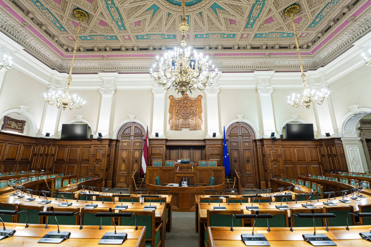 Tukša Latvijas Saeimas sēžu zāle ar koka deputātu soliem puslokā, prezidija tribīni centrā, kokgriezuma Latvijas ģerboni, Latvijas un Eiropas Savienības karogiem, grezni apgleznotiem griestiem un lielām lustrām. Kreisajā pusē redzama plāksne ar uzrakstu "LATVIJAS VALSTS SUVERĒNĀ VARA PIEDER LATVIJAS TAUTAI".