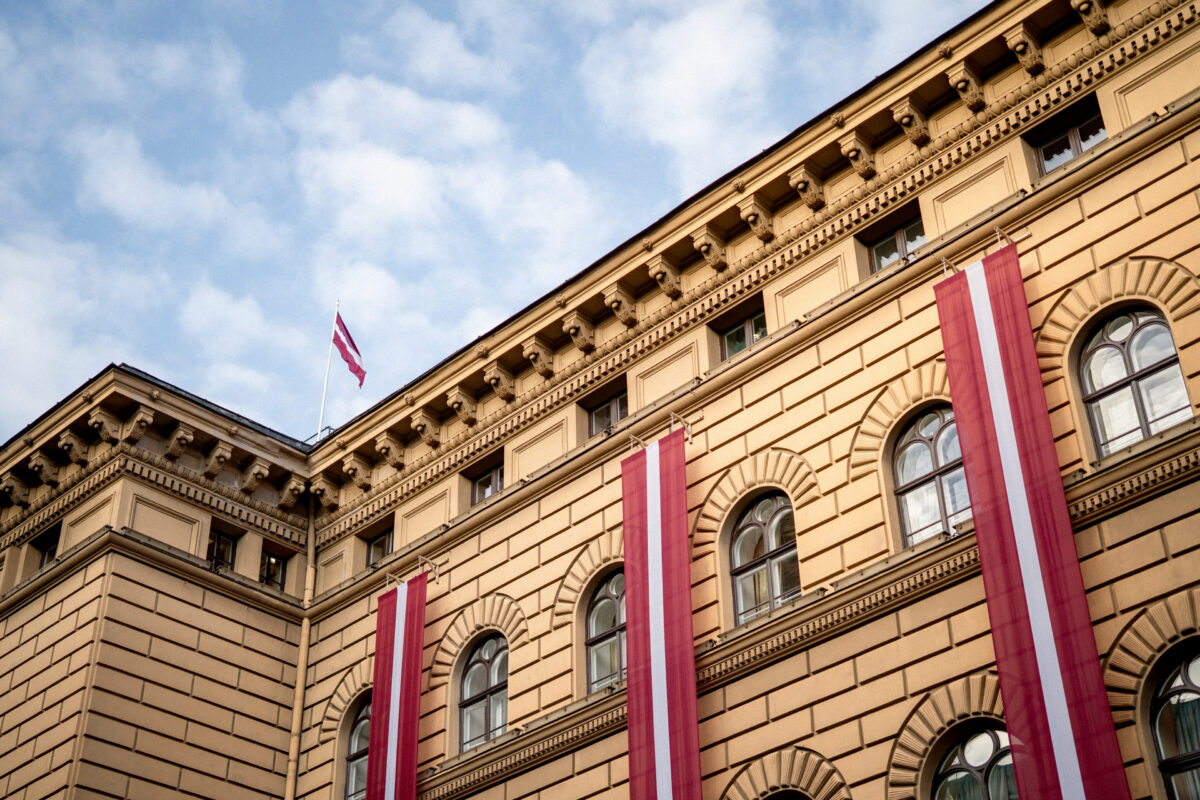 Saeimas ēkas smilškrāsas fasāde ar arkveida logiem, pie kuras vertikāli izkārti Latvijas valsts karogi, un uz jumta mastā plīvojošs karogs pret mākoņainām debesīm.
